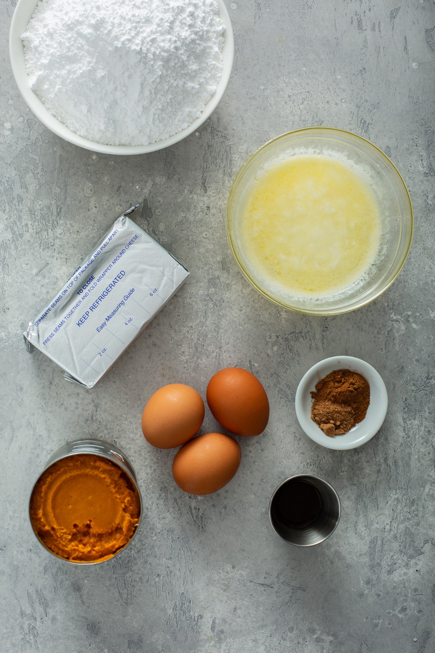 Ingredients for pumpkin gooey butter cake. Bowls of cake mix, melted butter, cinnamon, vanilla, three brown eggs, a can of pumpkin, and 