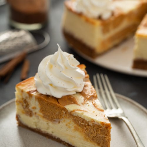 A slice of swirled pumpkin cheesecake on a plate with a fork. The rest of the cheesecake is in the background.