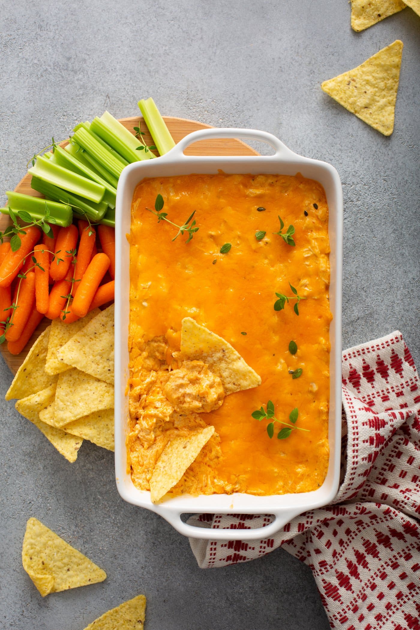 A casserole dish of instant pot buffalo chicken dip. There are two chips that have been dipped into the cheesy dip. Chips, carrots, and celery are next to the dip and a red and white checked tea towel is next the the dish.