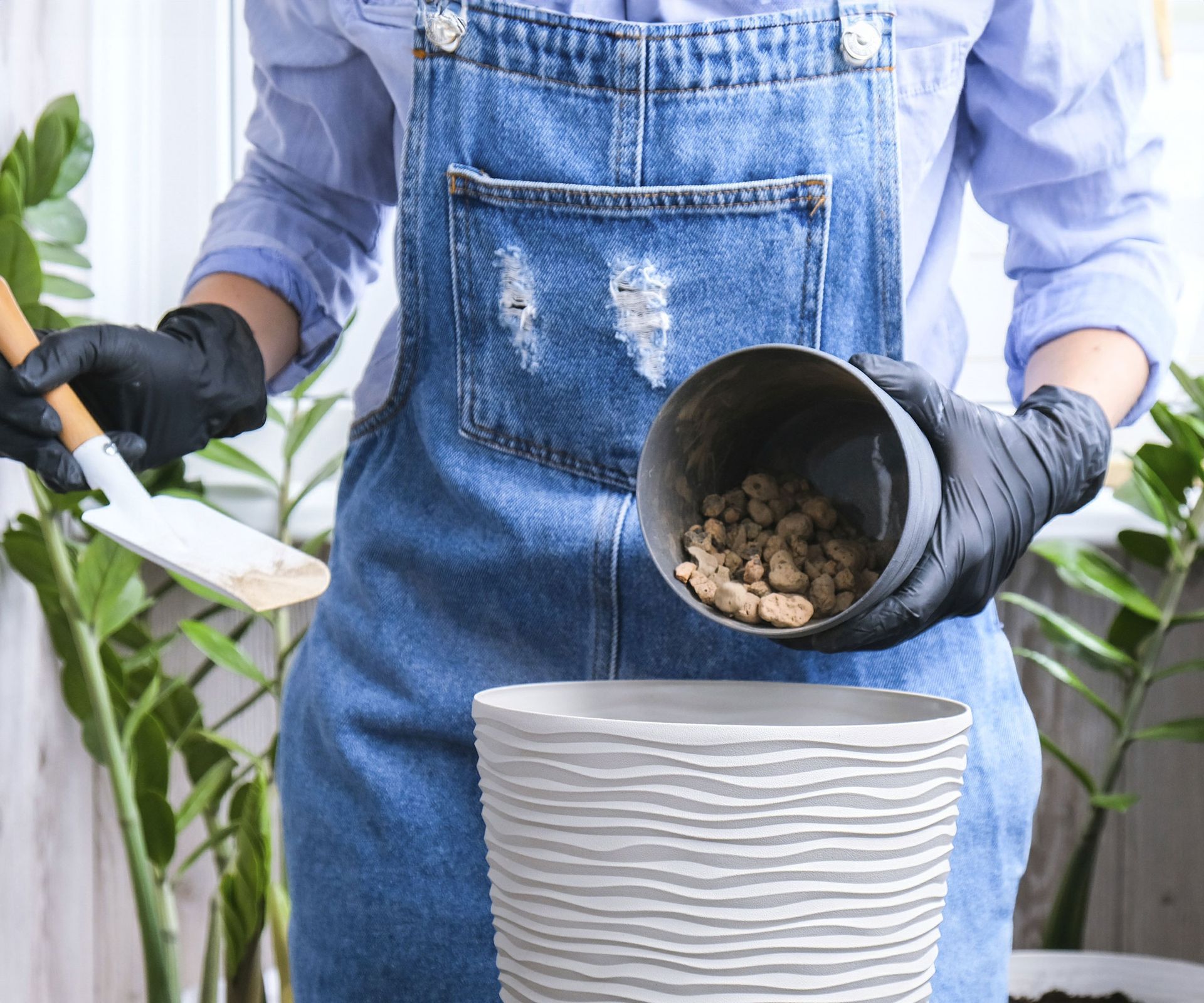person wearing blue denim overalls holding plastic plant pot filled with what looks like gravel