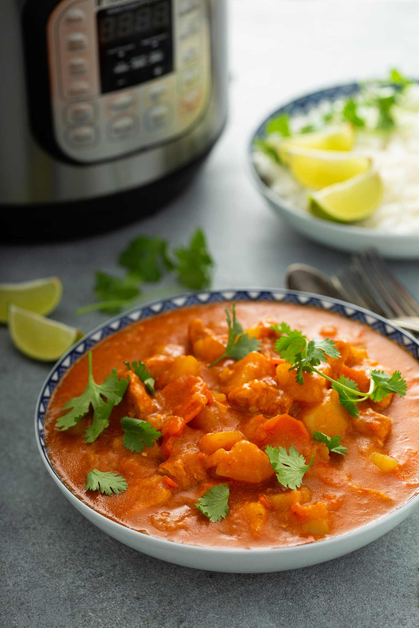 A bowl of chicken curry with chunks of chicken, potatoes and carrots in a curry sauce. Fresh parsley leaves are sprinkled on top and there is an instant pot, and lime wedges in the background.