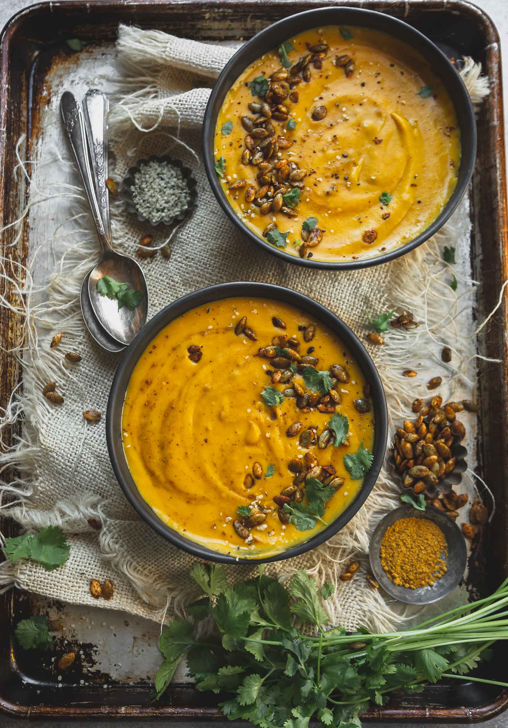 Two bowls of butternut squash soup. The soup is sprinkled with cilantro and hemp seeds. There is a spoon next to the bowls of soup.