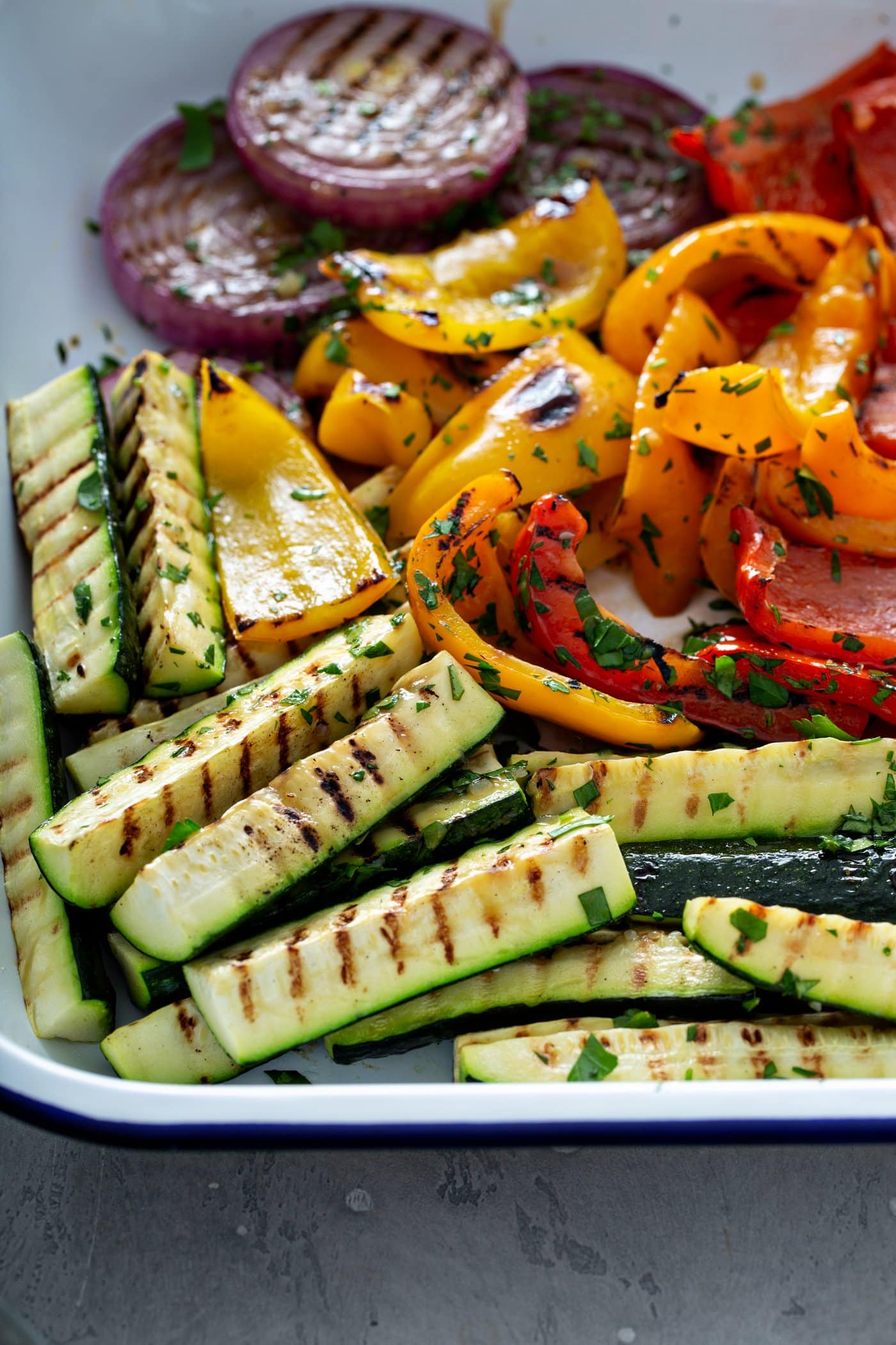 A plate full of grilled veggies. The zucchini wedges, red onion slices and red and yellow bell peppers have grill marks on them and they are sprinkled with a few parsley leaves.