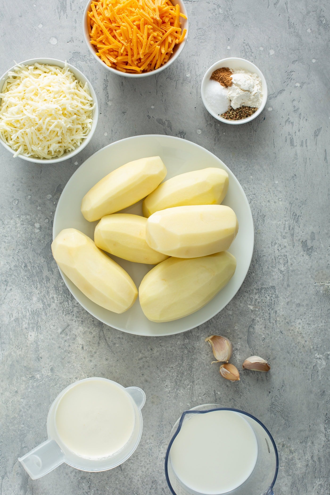 A bowl of six peeled potatoes. There are small containers of grated cheese, 