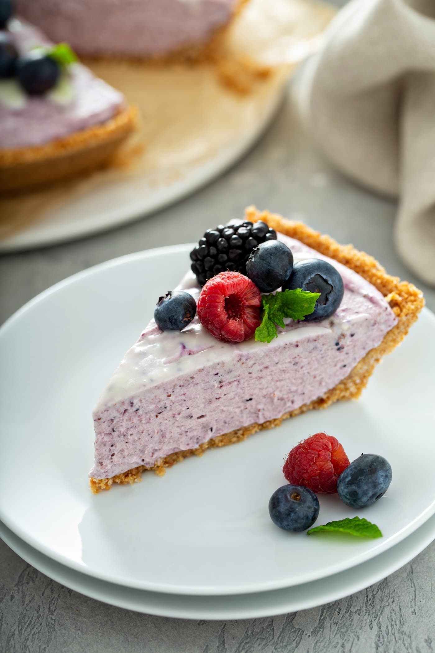 A slice of white chocolate berry pie on a dessert plate. There are a few mixed berries and mint leaves on the plate and the pie slice is topped with raspberries, blue berries and black berries. A mint leaf is next to the berries and a white chocolate sauce is drizzled on top of the pie. A pie plate with a whole pie and a napkin are in the background.