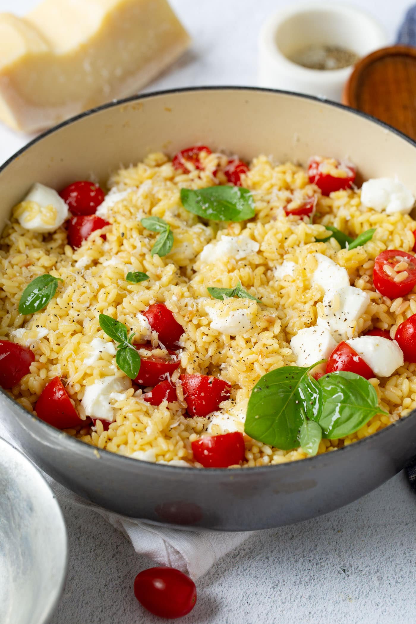 Caprese orzo salad in a larch serving bowl. There are bright red tomatoes, fresh green basil and chunks of mozzarella cheese. A few tomatoes, a brick of parmesan cheese, and a container of salt are on the table by the salad.