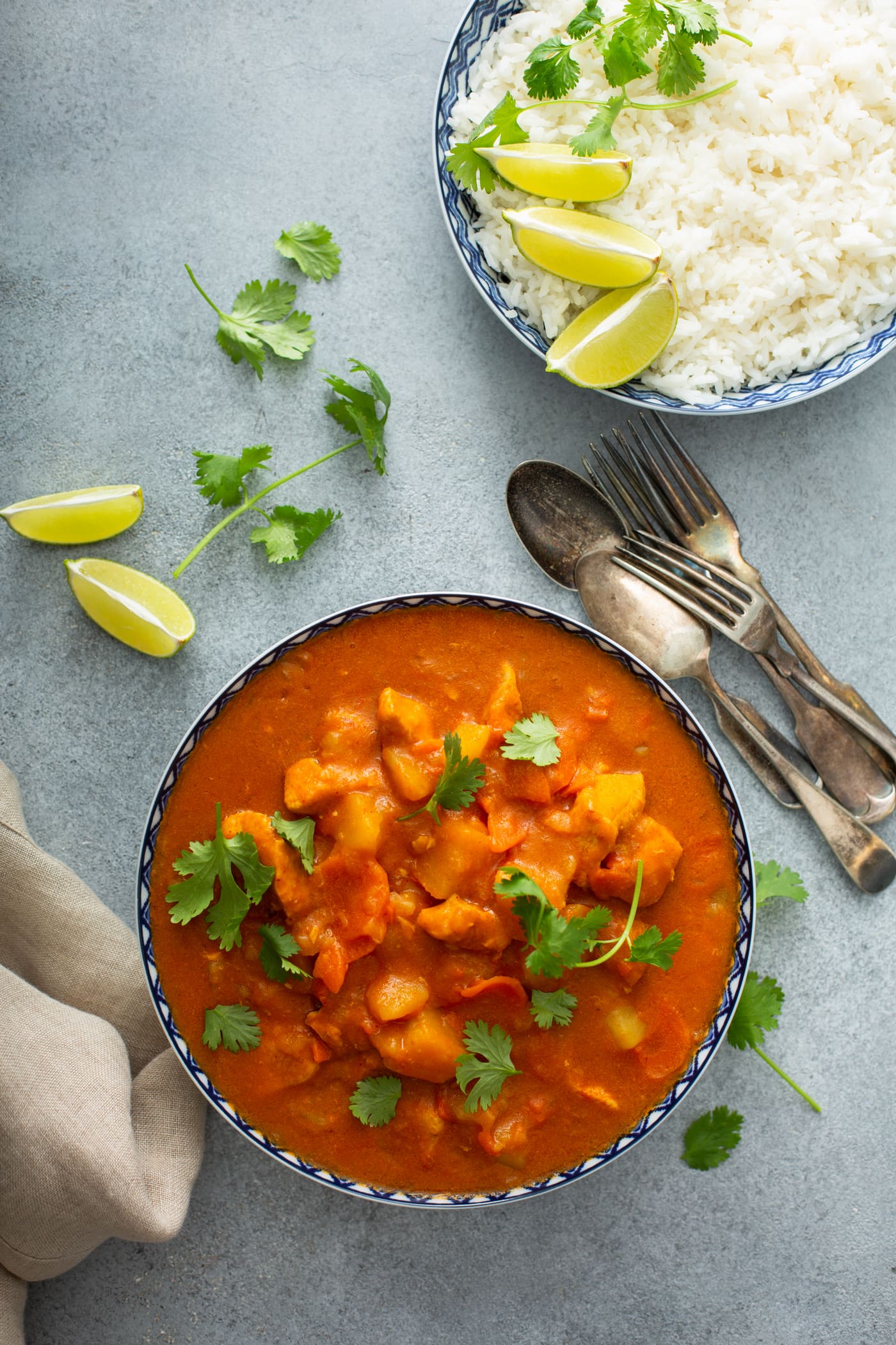 A bowl of instant pot chicken curry with chunks of chicken, carrots and potatoes. Fresh parsley leaves are scattered on top and on the table. there are lime wedges and silver ware in the background.