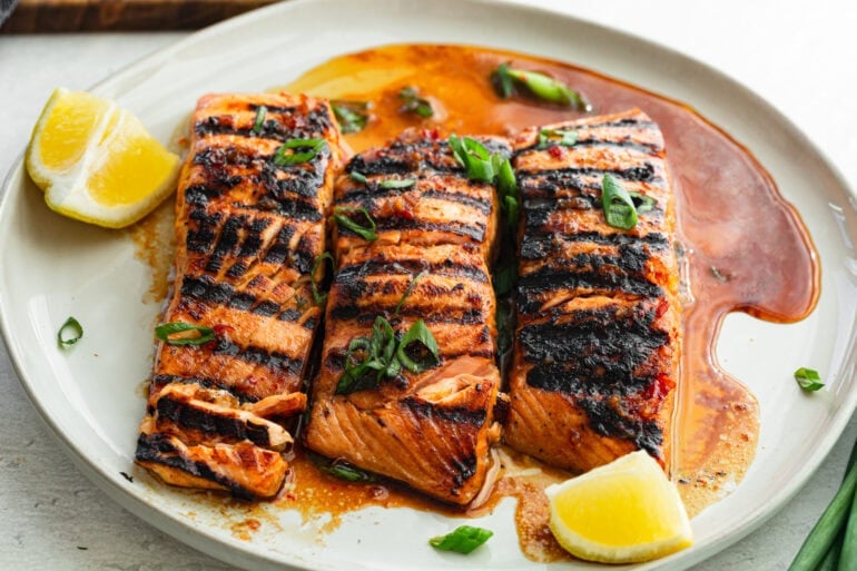 three marinated grilled salmon filets sitting on a white plate with a slice of lemon next to it and chopped green onions