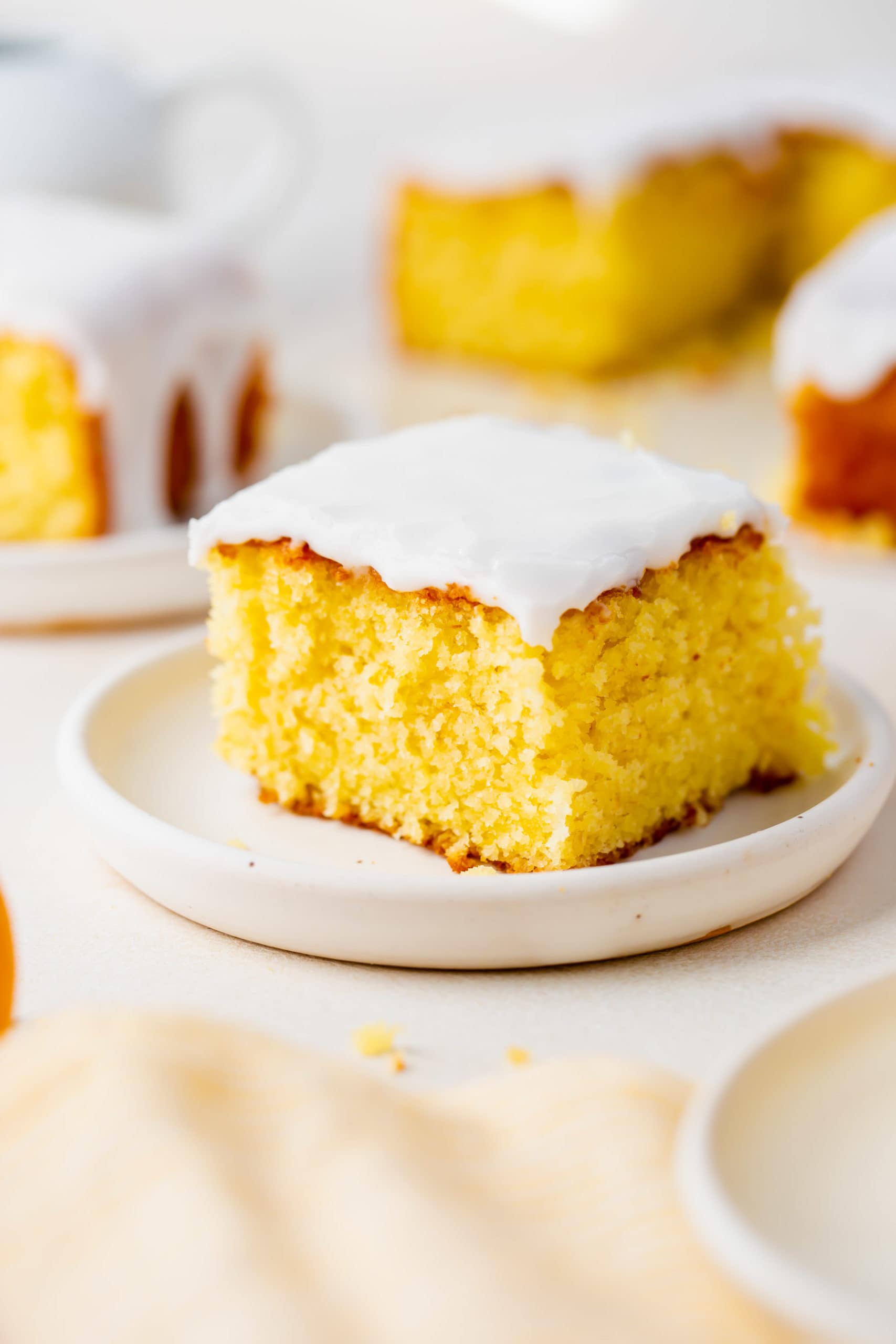 a photo of one piece of lemon cake topped with white frosting and sitting on a white plate. Several servings of cake are in the background.