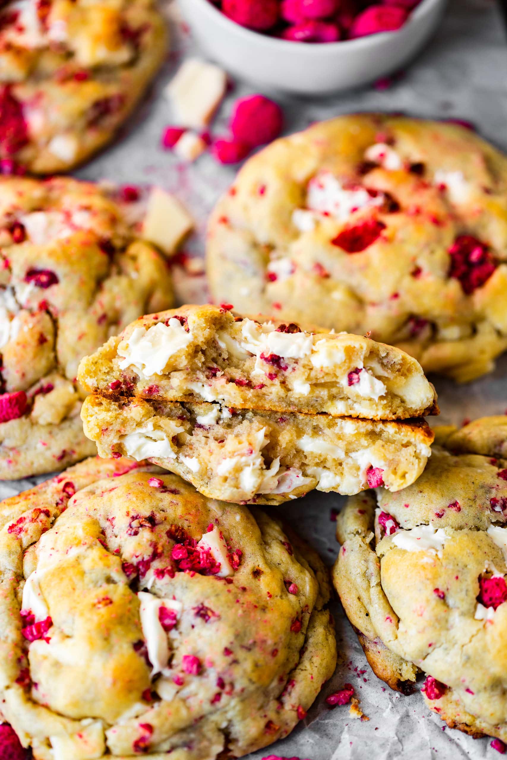 a photo of a thick white chocolate raspberry cookie that has been broken in half and then stacked on each other so you can see the inside of the cookie. It is sitting on a pile of other baked cookies.