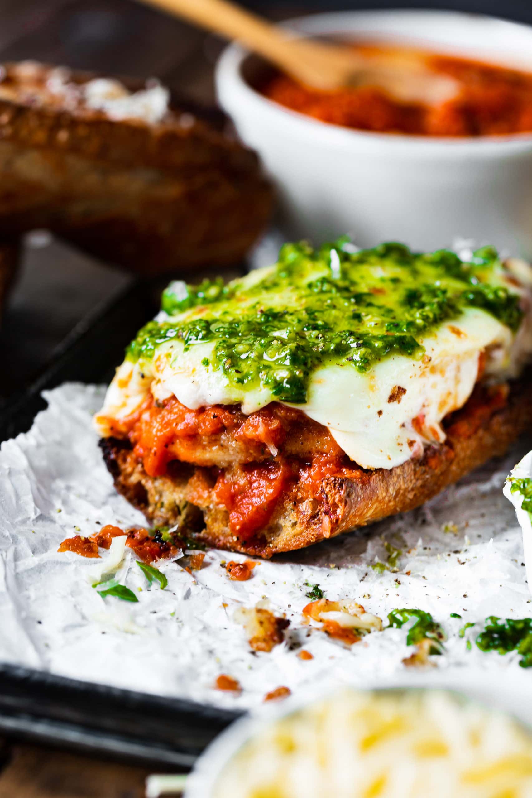 a photo of the bottom half of a chicken parmesan sandwich topped with melted mozzarella cheese and bright green pesto.