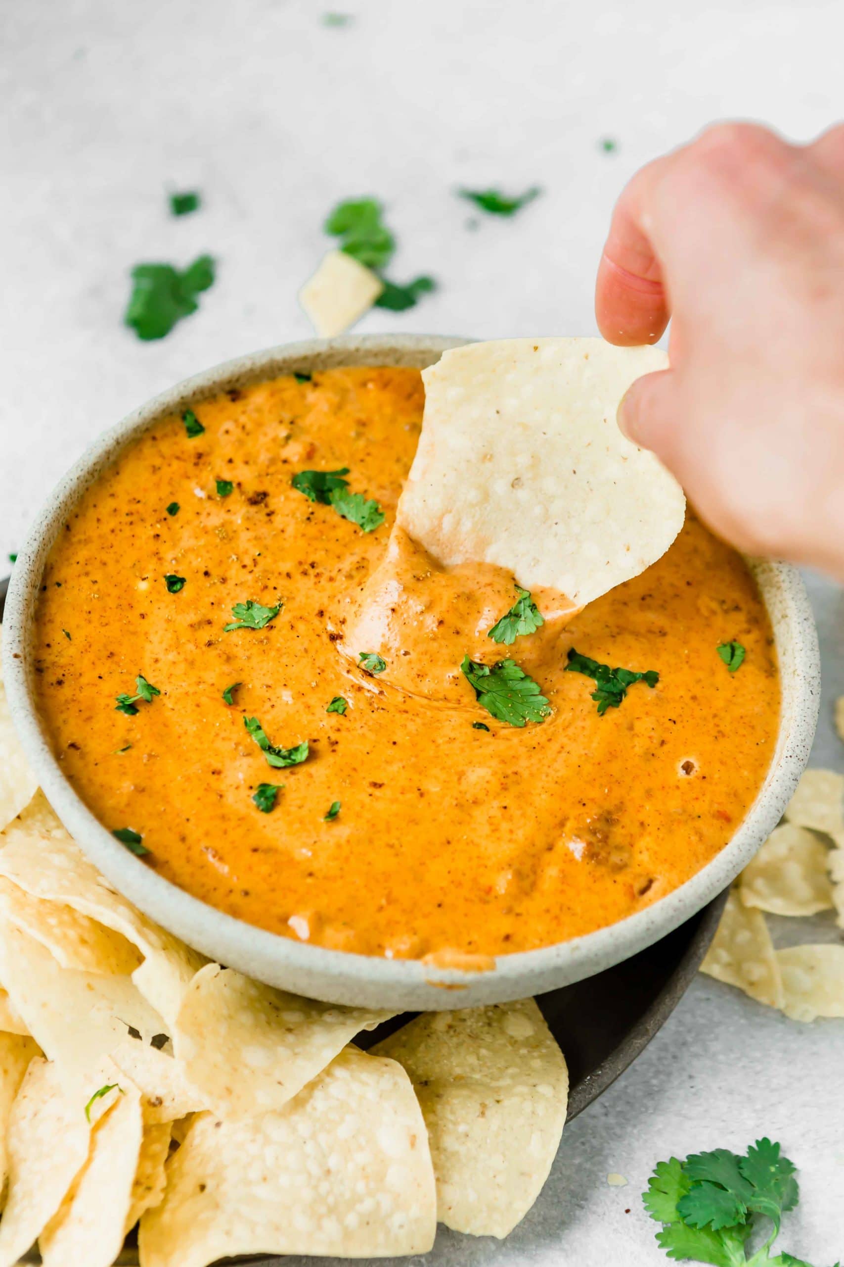 A bowl of chilis queso dip. It is cheesy and thick with a few parsley flakes scattered on top. A tortilla chip is being dipped into the cheese dip.