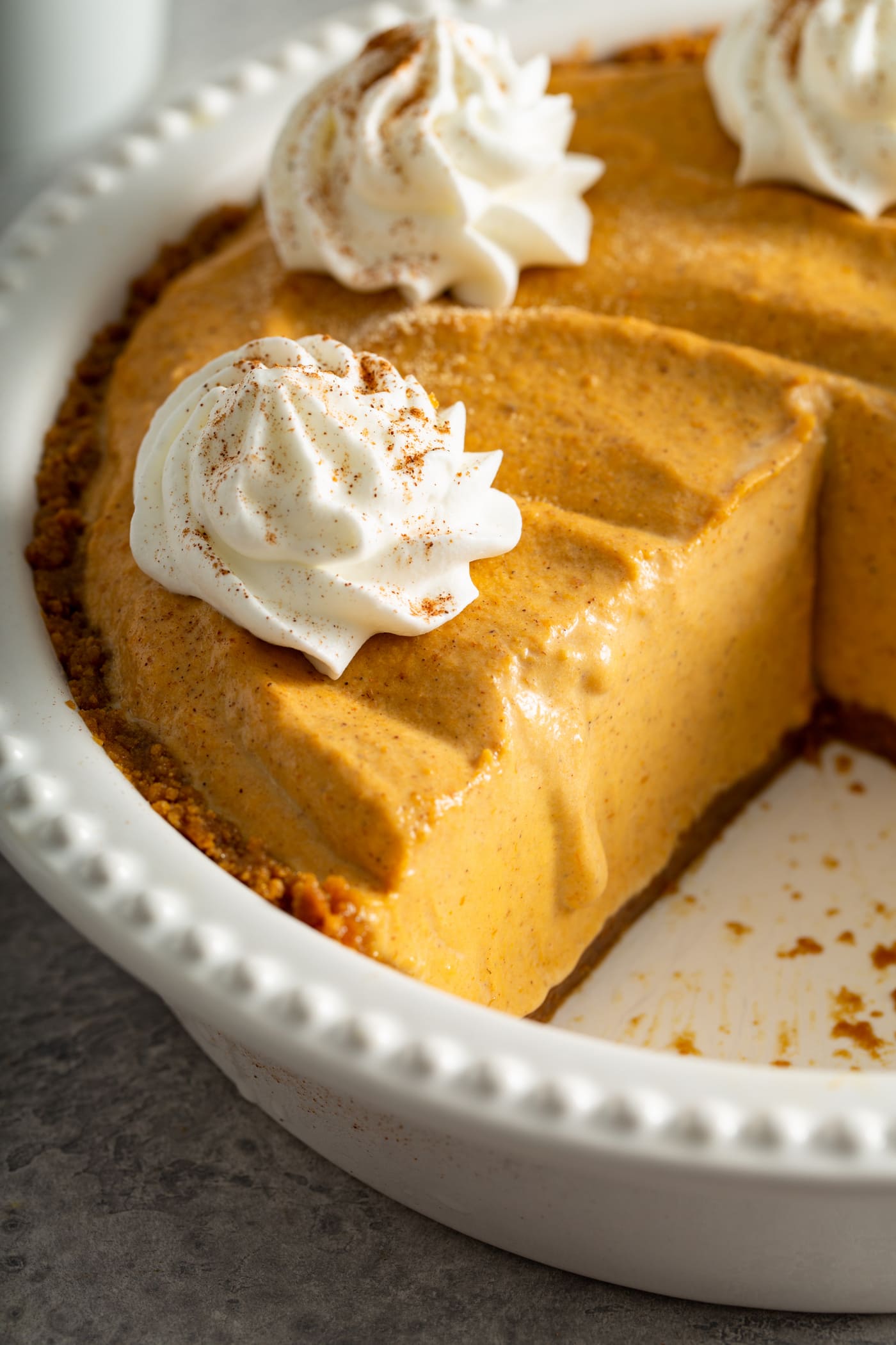 A pumpkin ice cream pie in a white pie dish with a slice removed. The pie has dollops of whipped cream sprinkled with cinnamon on top.