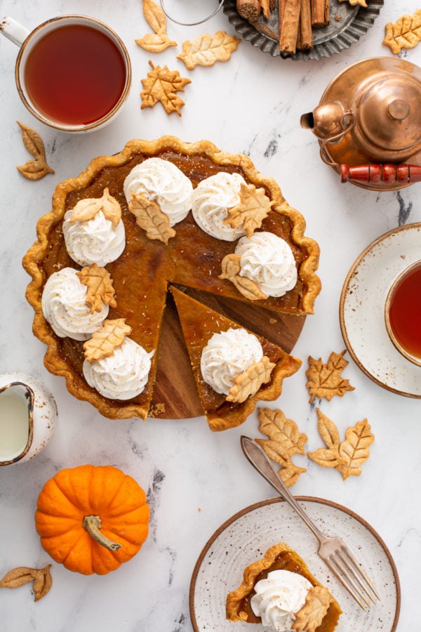 A photo of a pumpkin pie topped with piped whipped cream and stamped pie crust leaves taken from above with a slice pulled out.