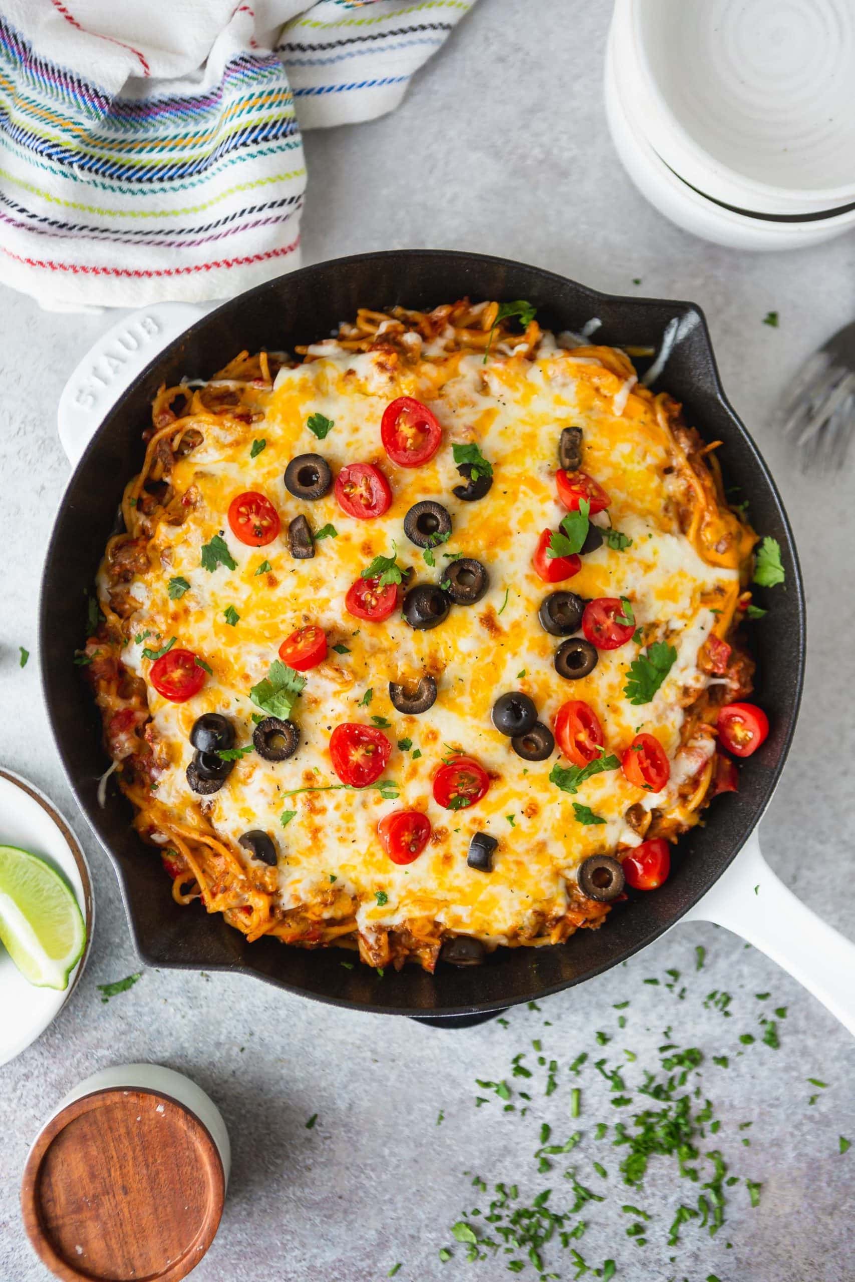 A cast iron skillet filled with cheesy ground beef taco spaghetti. It is topped with cheese, olives, green onion, tomatoes and cilantro.