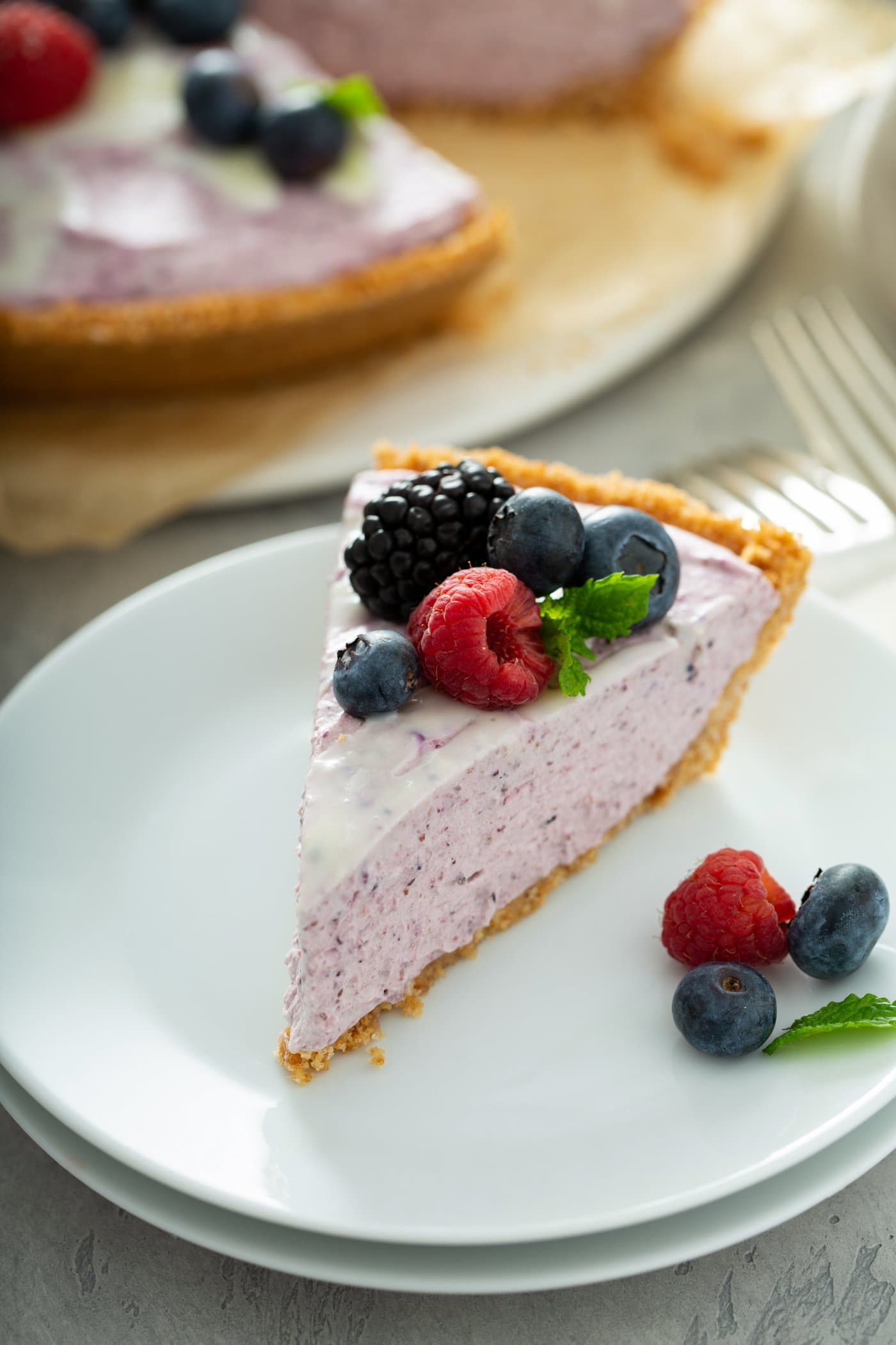 A white dessert plate with a slice of white chocolate berry pie. The pie is pink in color and you can see bits of pureed berries in the filling. the pie has raspberries, blue berries and black berries and a mint leaf on top. There are berries on the plate and a white chocolate berry pie and a couple of forks in the background
