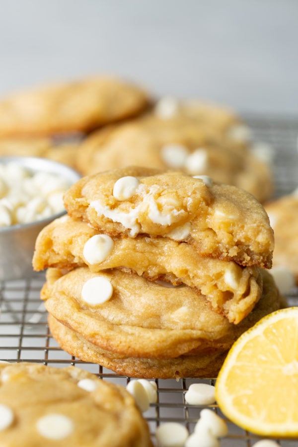 a photo of a stack of golden lemon cheesecake cookies full of white chocolate chips with the top cookie broken in half with the insides of cookie showing.