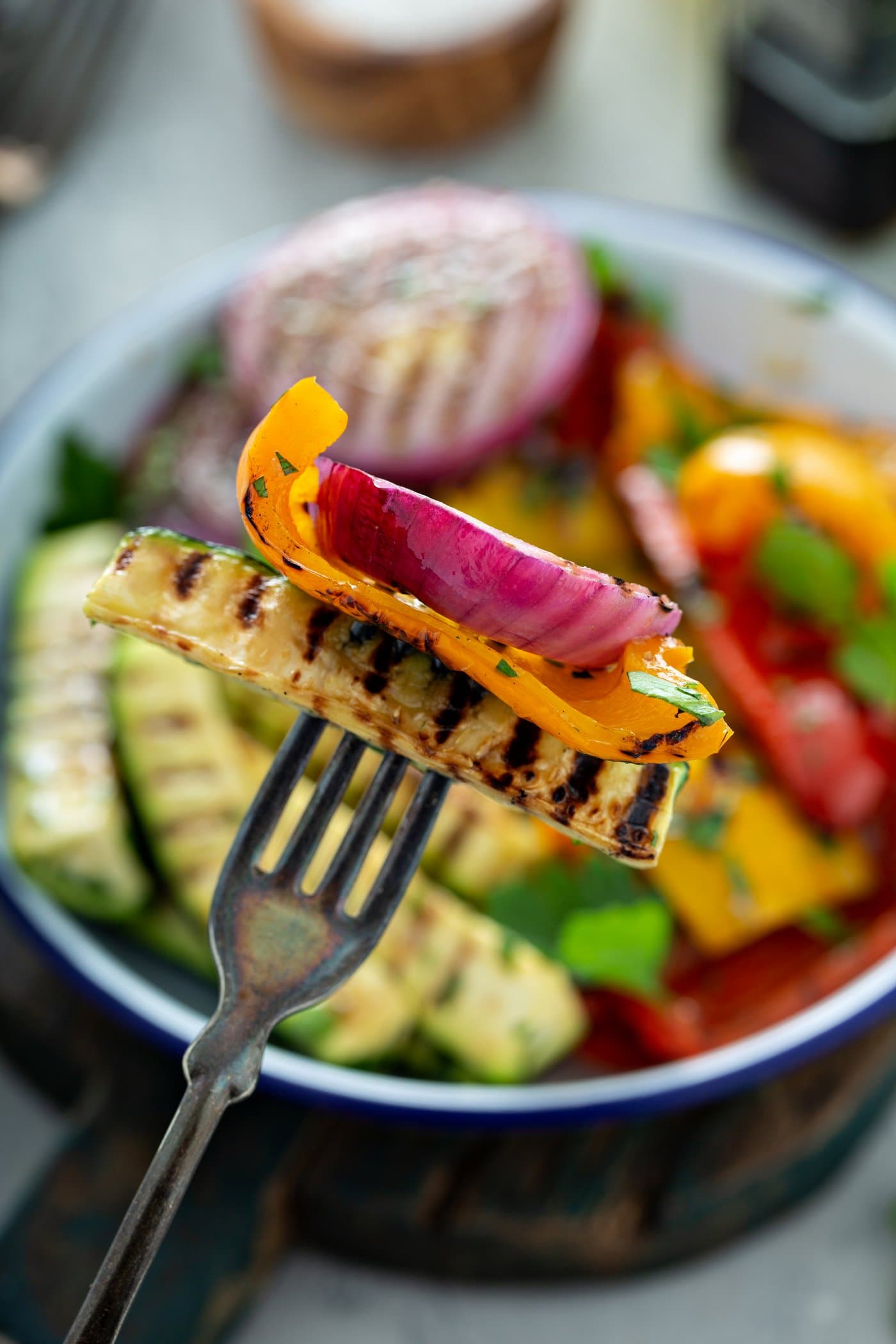 Grilled zucchini, yellow bell pepper, and red onion pierced with a fork. A serving dish of grilled veggies is in the background.