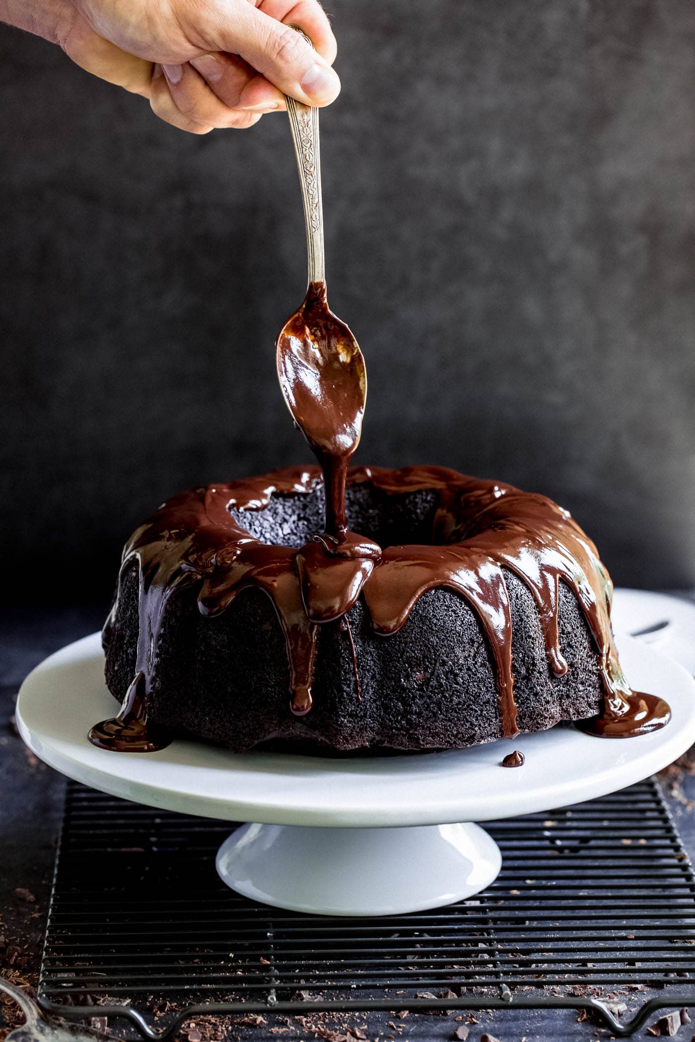 A spoon with thick chocolate glaze drizzling over the top of a zucchini chocolate bundt cake which is on a white cake plate. 