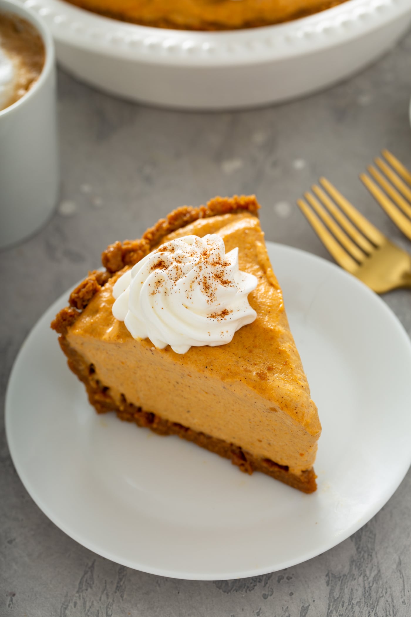 A slice of pumpkin ice cream pie on a white dessert plate. The pie is topped with a dollop of whipped cream with cinnamon sprinkled on top. Two forks are on the table next to the plate.