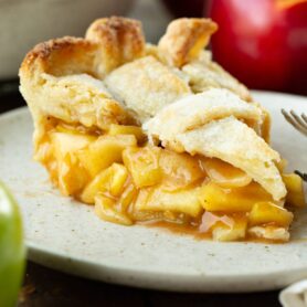 a photo of a slice of homemade apple pie sitting on a small dessert plate. The crust is golden and a lattice top and the filling is gooey with cinnamon and perfectly soft apples.