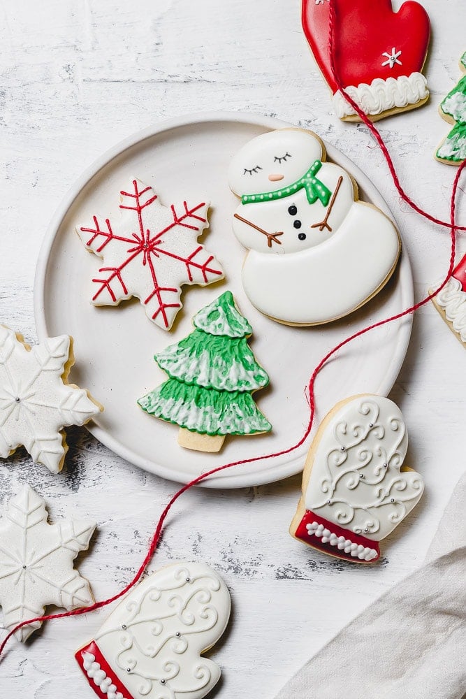 A plate with Christmas sugar cookies decorated with royal icing. There is a snowman with a green scarf, a white mitten with red trim, a green tree with white snow accents, and a white star with red accents. A red ribbon is draped over the edge of the plate.