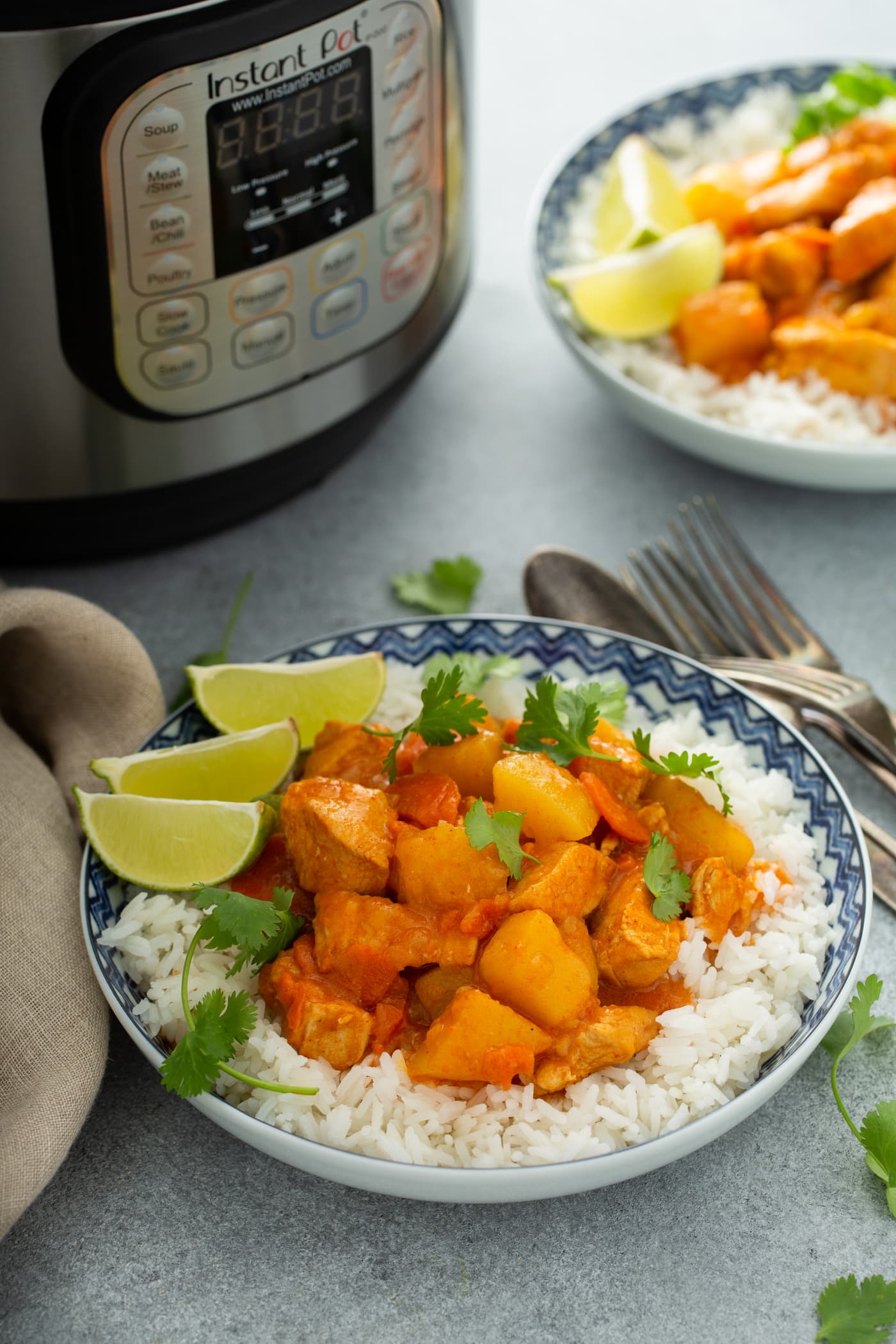 Two bowls of white rice topped with chicken curry. There are lime wedges on the edge of the bowls and fresh parsley sprinkled on top of the curry. An instant pot is in the background and forks and spoons are next to the curry.