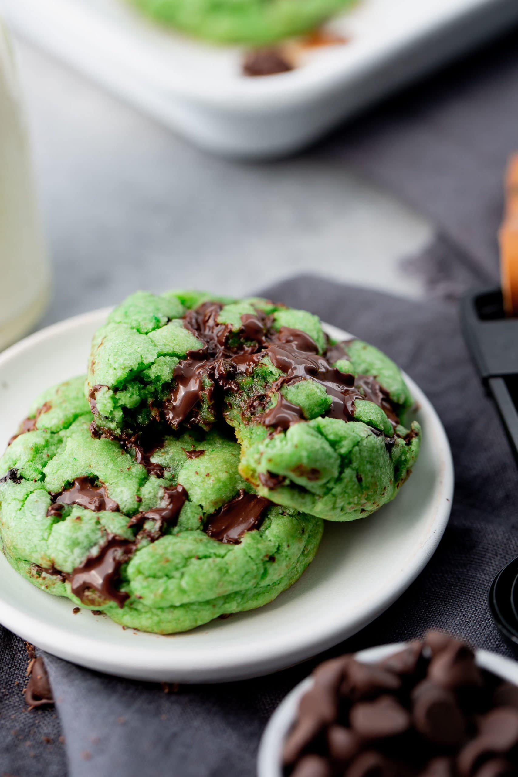 a photo of two green mint chocolate chip cookies on a white small plate. one cookie has a bite taken out of it.