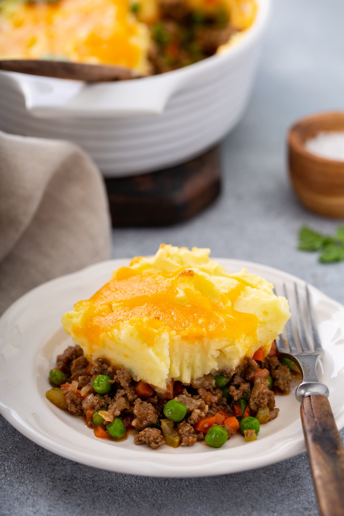 A serving of shepherd's pie with carrots, peas and ground beef for filling, topped with mashed potatoes and melted cheese. There is a fork resting on the plate and a napkin and a small container of salt next to it.