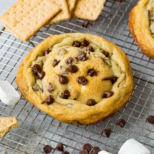 A s'mores stuffed chocolate chip cookie. There are graham crackers, chocolate chips and marshmallows in the background.