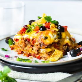 a photo of a piece of taco casserole sitting on a saucer topped with fritos, sliced olives, diced tomatoes, and chopped fresh cilantro.