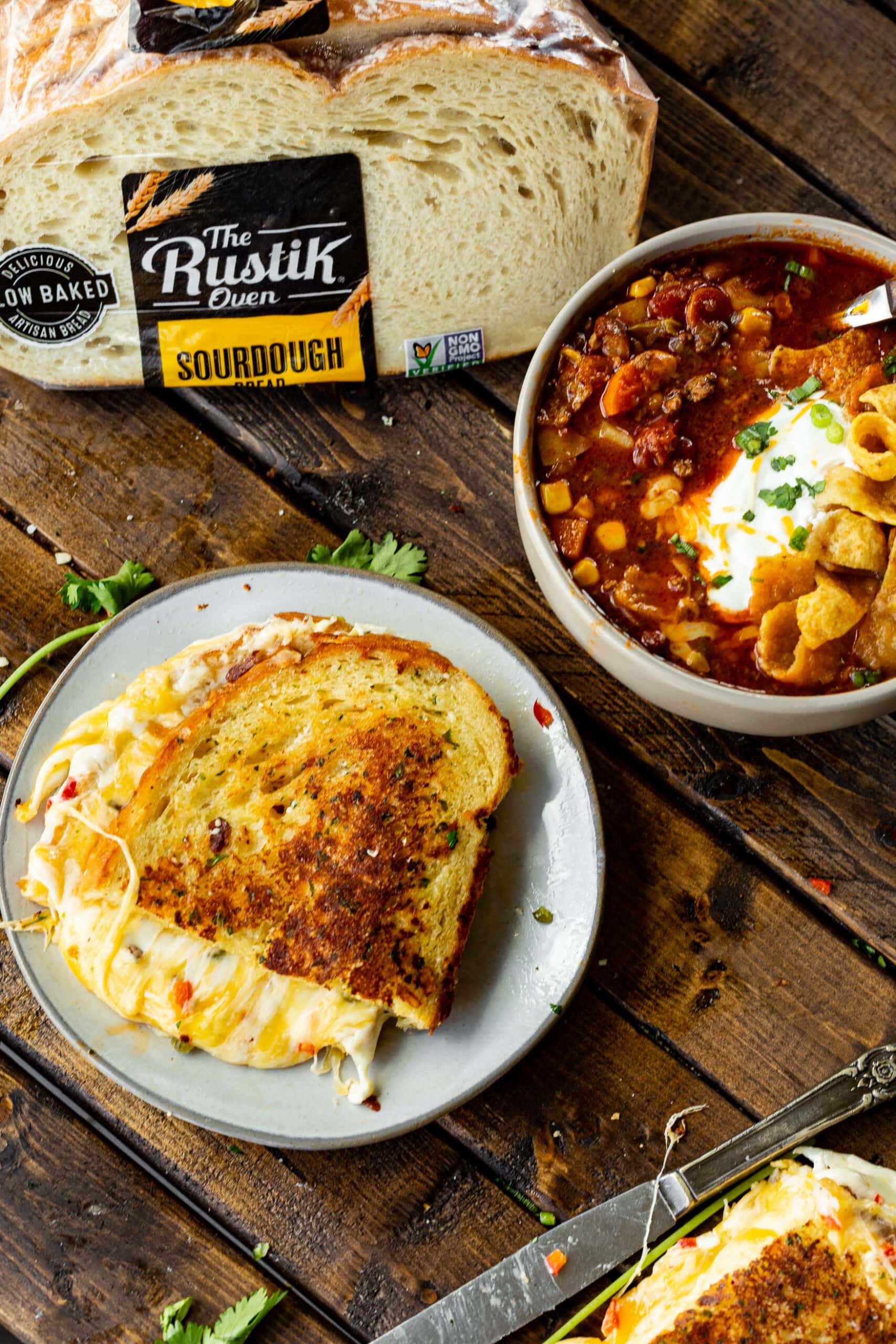 a photo of a half of a golden grilled cheese sandwich with melty cheese coming out all of the sides. The sandwich is sitting on a small plate and is sitting next to a bowl of taco stew and a bag of sourdough bread.