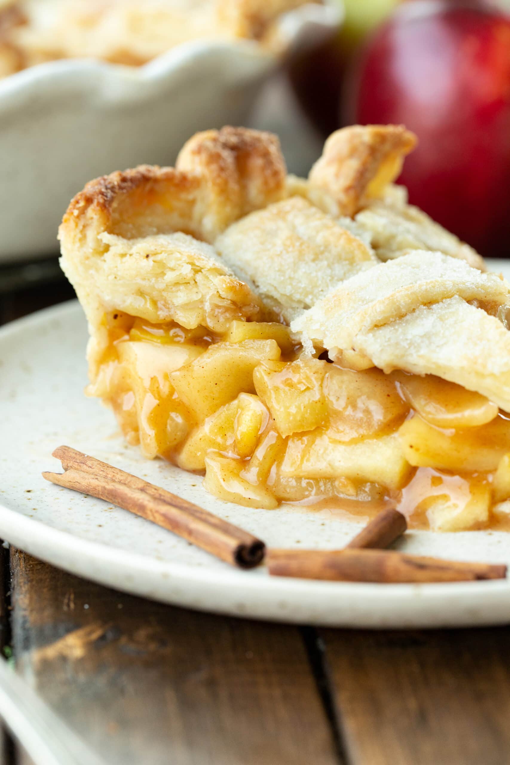a photo of a slice of homemade apple pie with a lattice top sprinkled with sugar. There are a few cinnamon sticks scattered in front of the piece of pie.