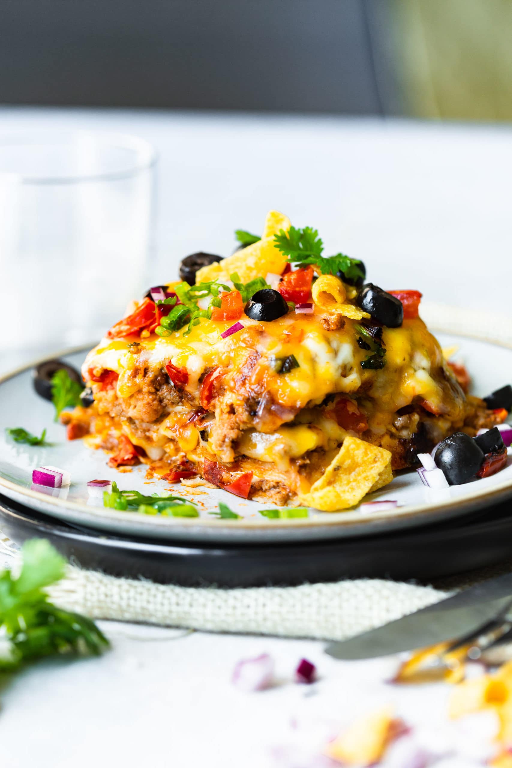 a photo of a piece of taco casserole sitting on a saucer topped with fritos, sliced olives, diced tomatoes, and chopped fresh cilantro.