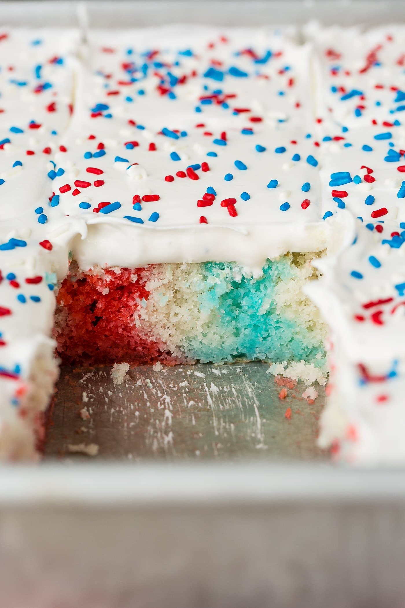Red, white and blue cake with a slice removed exposing the red, white and blue inside of the cake. The cake has white frosting and red, white and blue sprinkles on top.