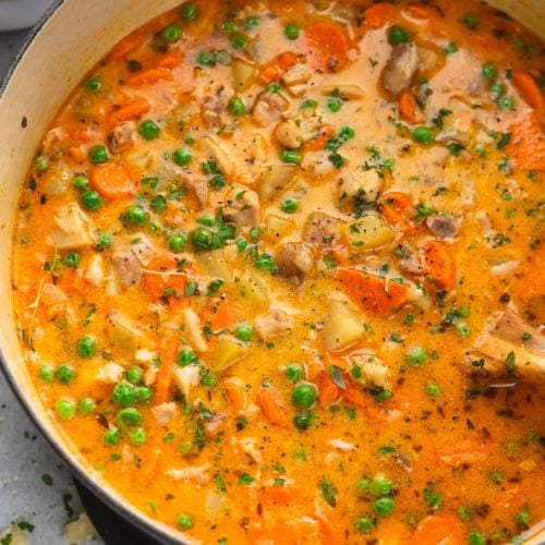 A pot of slow cooker chicken stew. There are bits of vegetables, chicken, and herbs in a reddish yellow gravy.