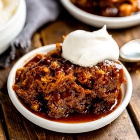 a photo of a serving of pumpkin lava cake in a small white saucer. the cake is sitting in a thick sauce that has seeping out of the cake and the cake is topped with whipped cream.