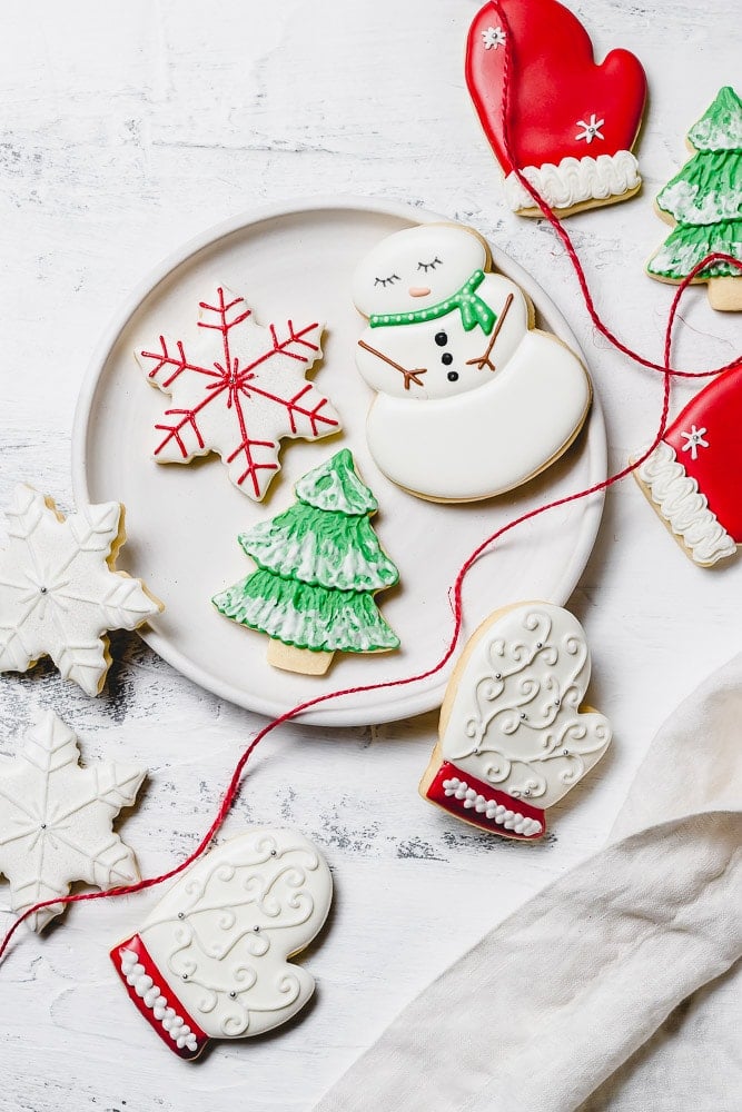 A plate of Christmas sugar cookies decorated with royal icing. There is a snowman with green scarf, a snowflake with red accents, a green tree with white snow, a white mitten with red trim, and white snowflakes