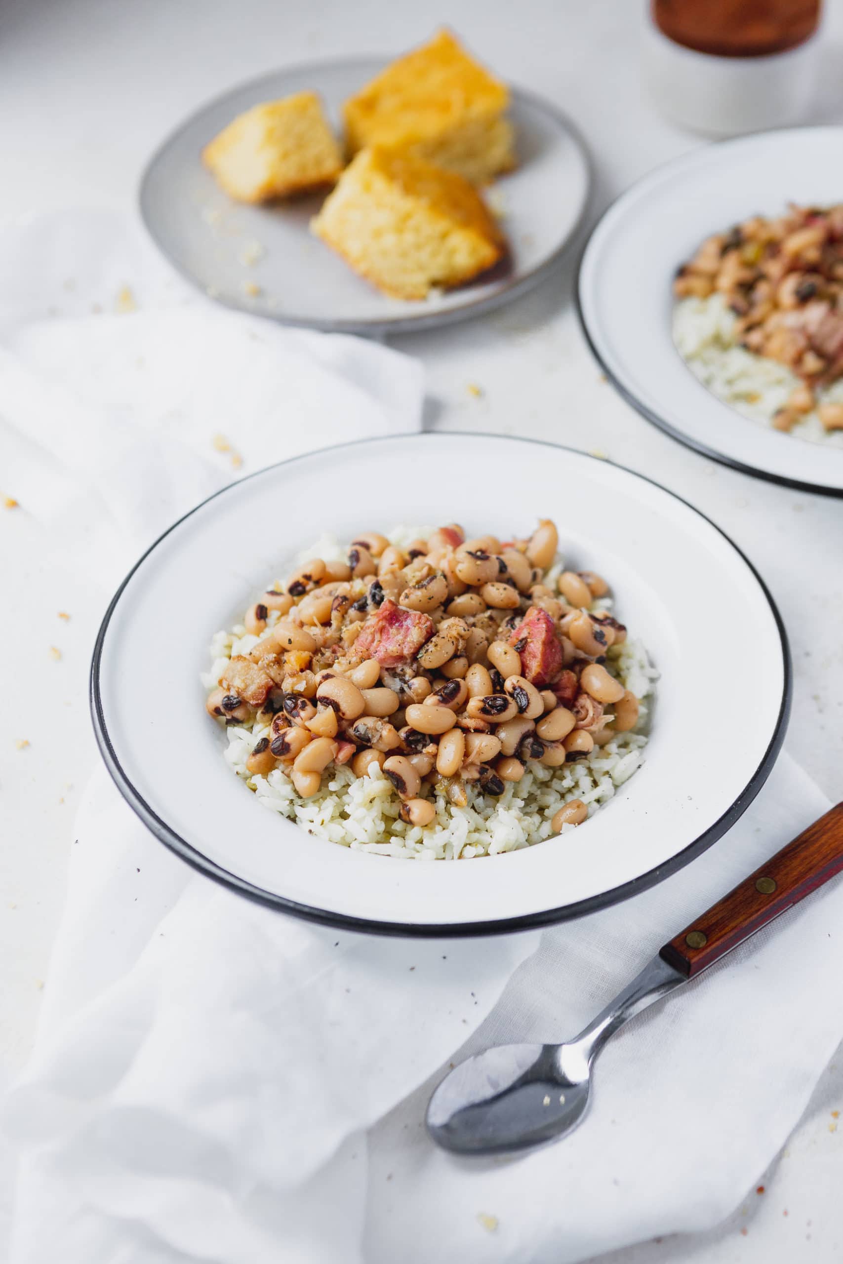 A plate of rice with southern instant pot black eyed peas on top. There is a spoon beside the plate and another plate of peas and a plate of corn bread in the background.
