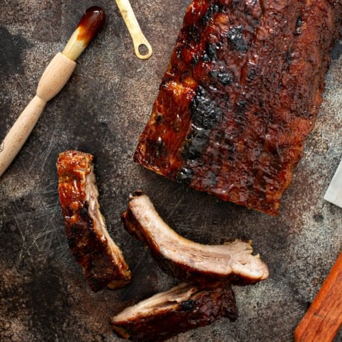 A rack of grilled ribs with three ribs sliced from the rack. A brush with glaze for the ribs is next to the rack of ribs.