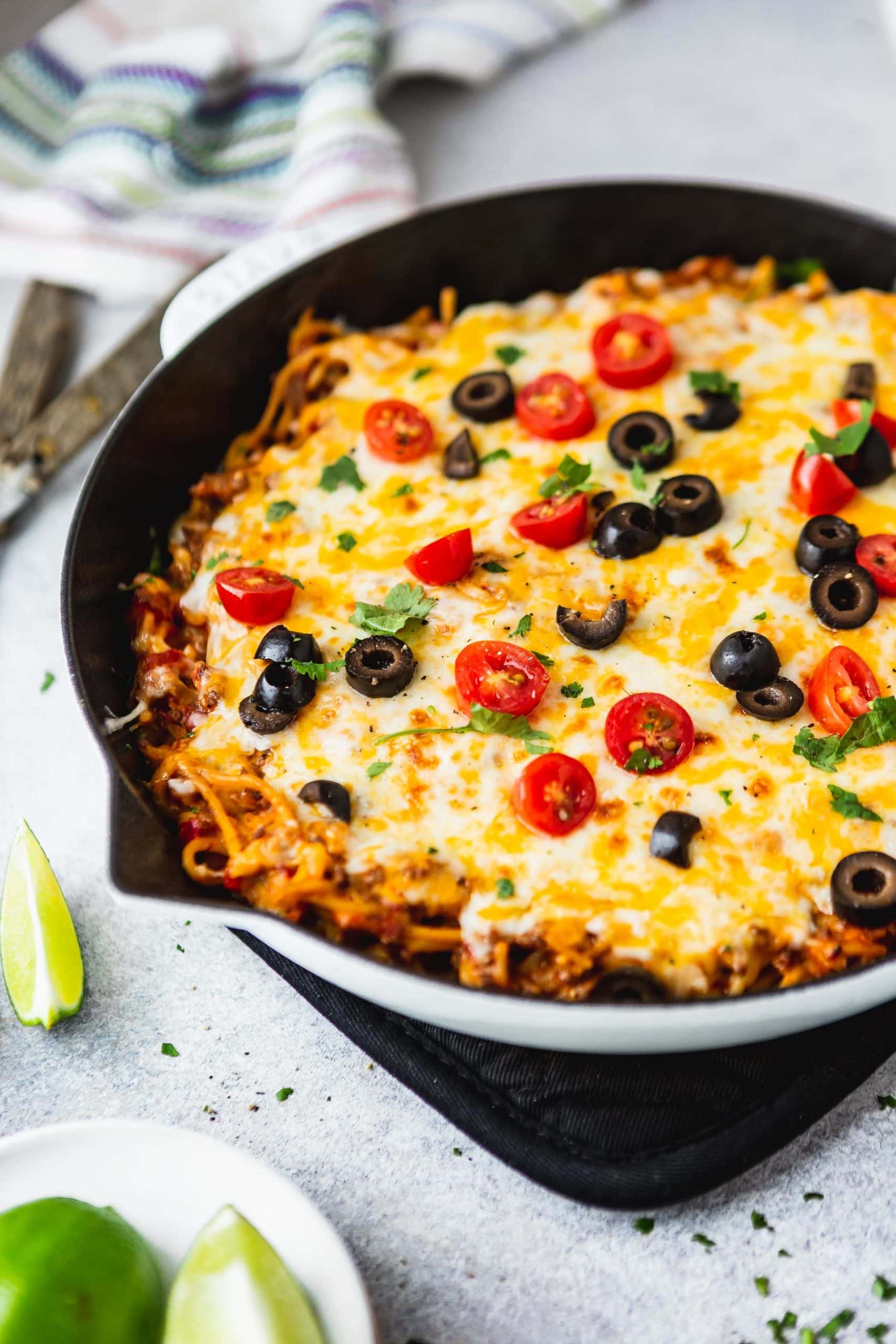 A cast iron skillet filled with cheesy ground beef taco spaghetti. There is melted cheese, olives, green onions, tomatoes and cilantro on top and a lime wedge is next to the skillet.