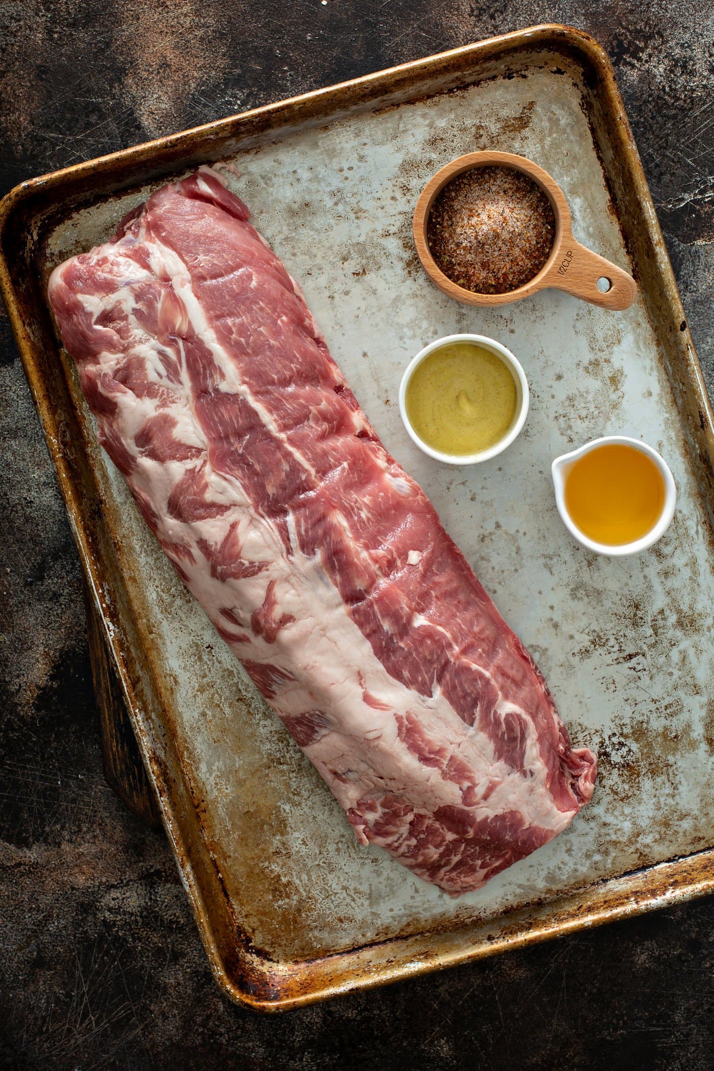 A rack of uncooked ribs on a baking pan. There are small containers of brown sugar, apple juice and mustard sitting next to the ribs.