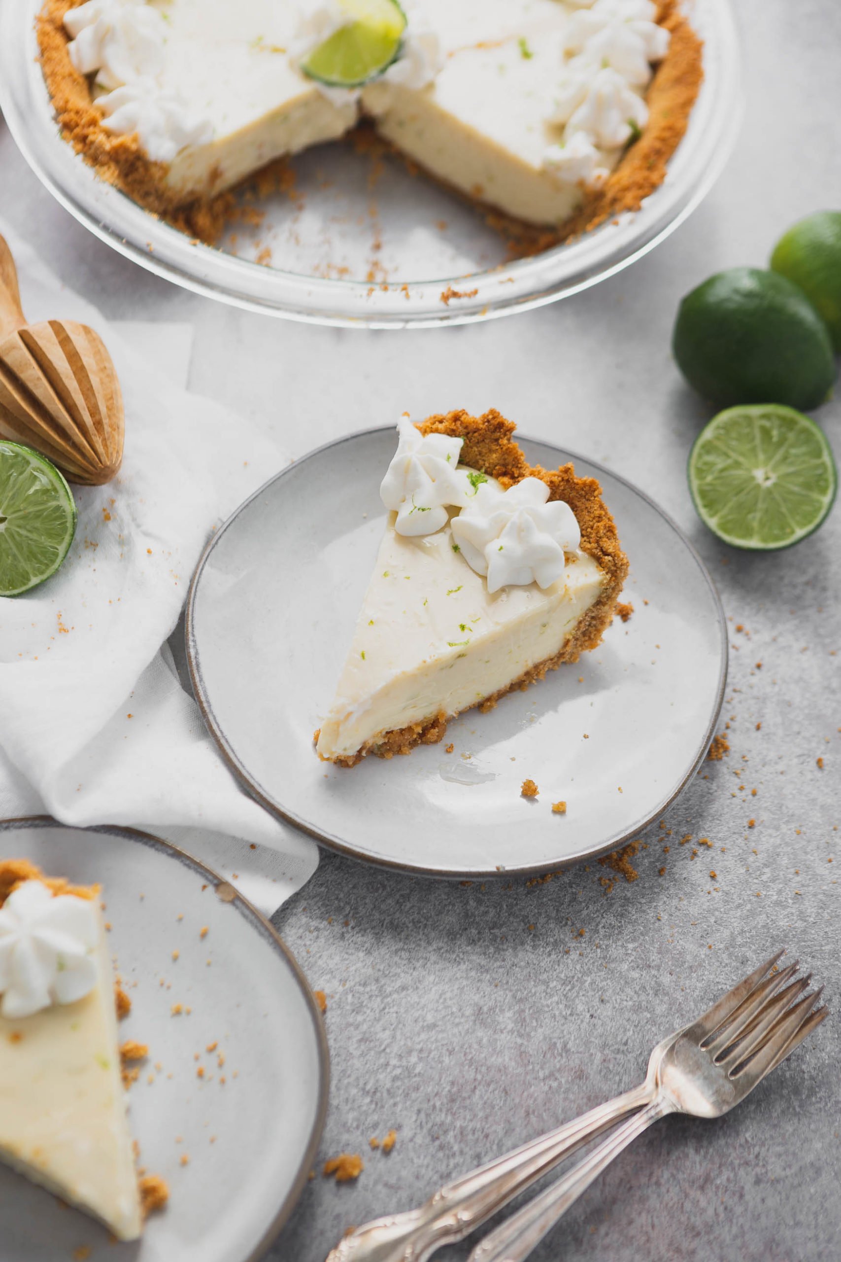 A slice of key lime pie on a white dessert plate. The graham cracker crust is filled with key lime filling and there are dollops of whipped cream on top. A lime that is cut in half is beside the plate and there is another dessert plate with a slice of pie on it.