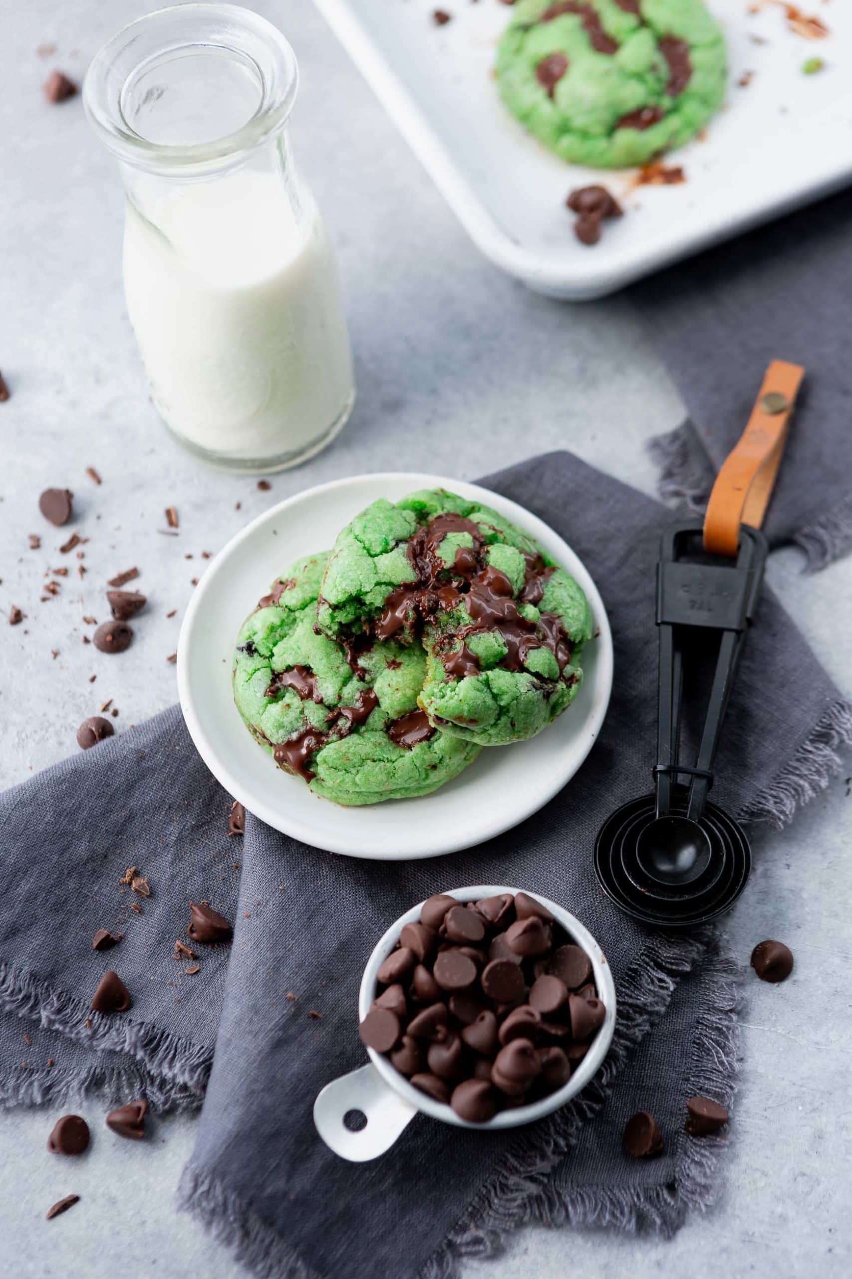 a photo of two green mint chocolate chip cookies on a white small plate. one cookie has a bite taken out of it. there is a glass of milk sitting next to them and a cup of chocolate chips.