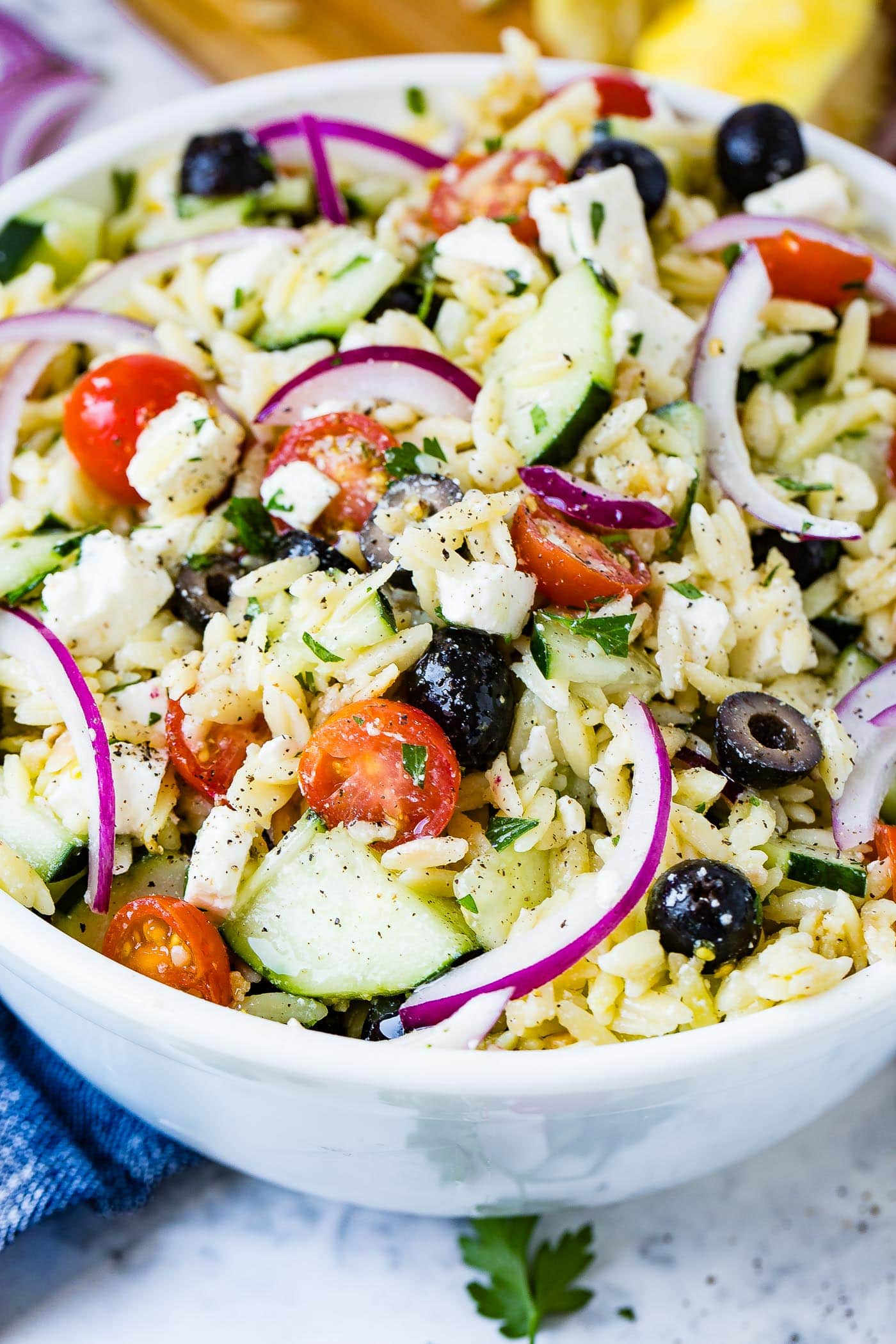 A white bowl of orzo pasta salad with red onion, cherry tomatoes, cucumbers, feta cheese and black olives.