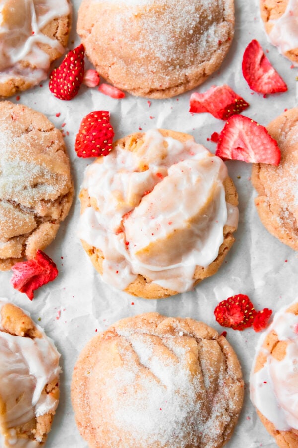 a photo of several baked strawberry cookies lined up on a white parchment paper. some of the cookies are stopped with a white glaze and other have been rolled in sugar.