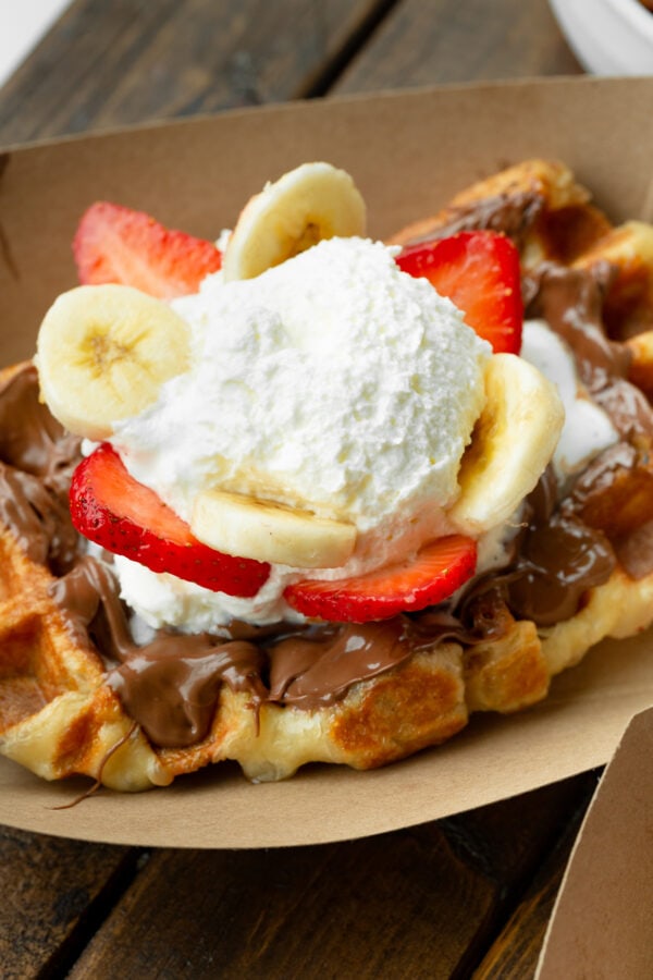 a photo of a golden liege waffle in a brown cardboard carton topped with nutella, sliced strawberries and bananas and whipped cream