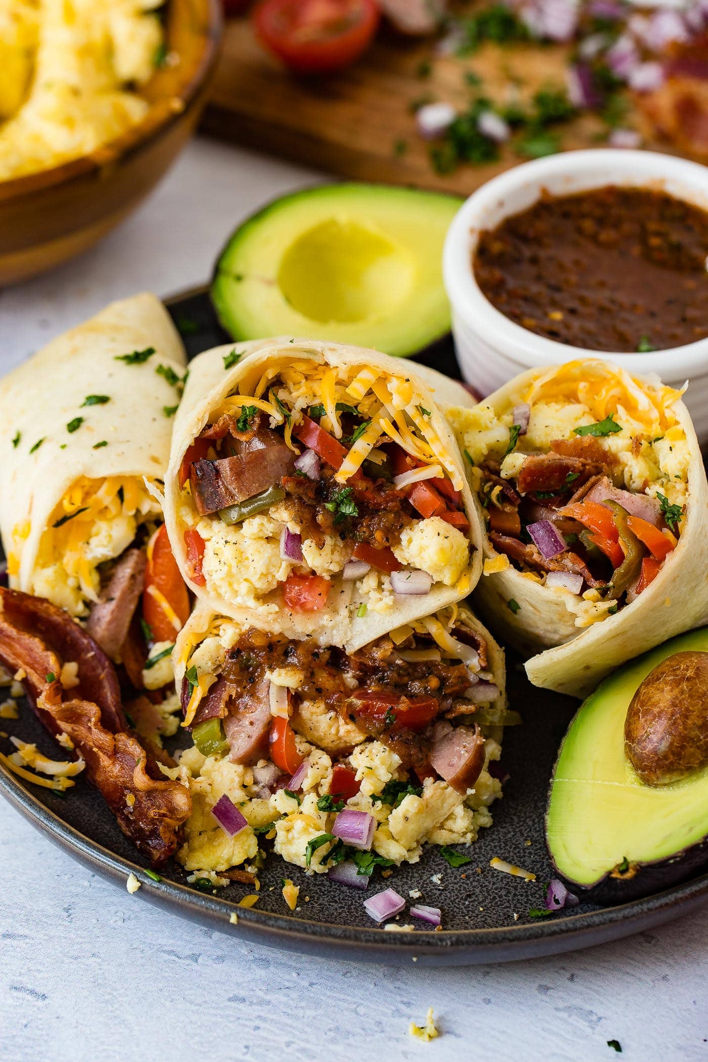 A plate with two apple chicken sausage burritos. The burrito has sausage, eggs, cheese. There is a bowl of salsa and an avocado that is cut in half next to the burritos.