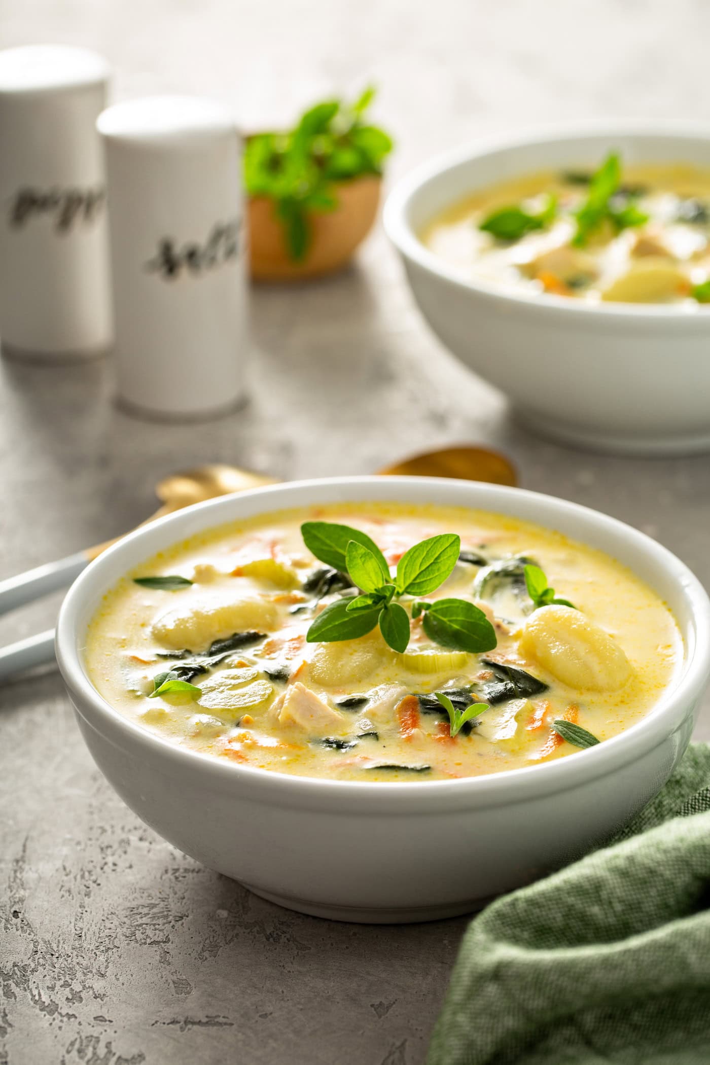 Two bowls of copycat Olive Garden chicken gnocchi soup. The soup has shredded carrots, gnocchi, celery and spinach in a creamy soup. Fresh herbs are sprinkled on top, and two soup spoons, salt and pepper shakers, and a green napkin are on the table next to the soup.