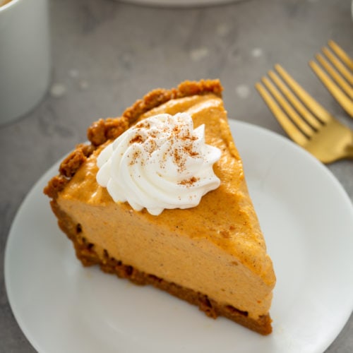 A slice of pumpkin ice cream pie on a white dessert plate. The pie is topped with a dollop of whipped cream with cinnamon sprinkled on top. Two forks are on the table next to the plate.